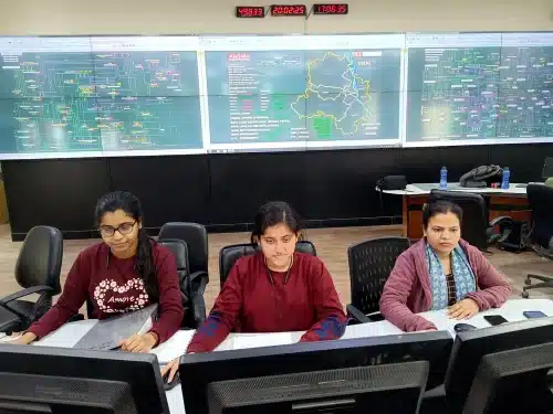 Three staff at a SCADA control room desk in Delhi's Balaji Estate, with large screens showing grid maps and real-time network data behind them