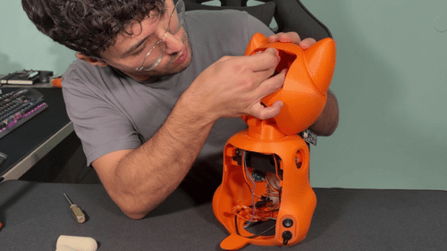 A maker assembling the 3D-printed orange fox-shaped enclosure of the Nuvi robot, with internal wiring and servos visible