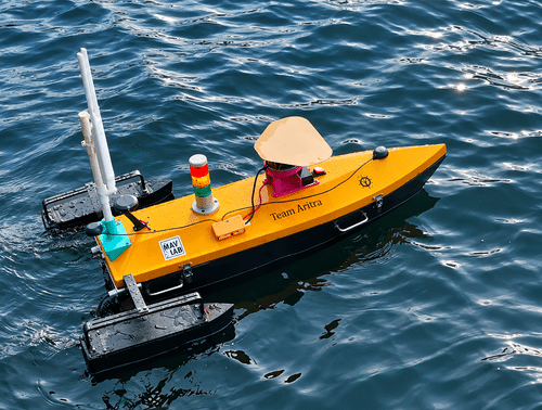 Team Aritra's autonomous surface vessel on the water, with GPS antennas, a lidar-style sensor dome, a signal light tower and black outrigger floats
