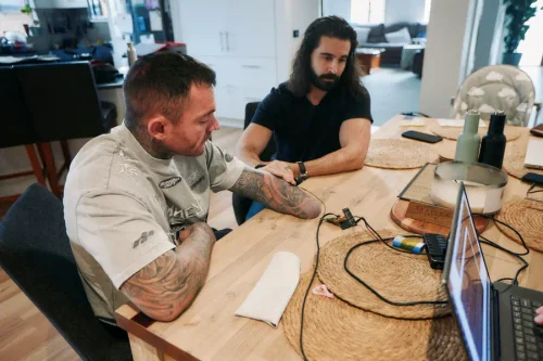 Two men seated at a table with wired electrode sensors and a laptop, reviewing muscle signal data