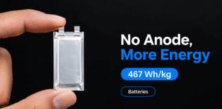 Thin silver anode-free battery pouch cell held between two fingers against a dark background.